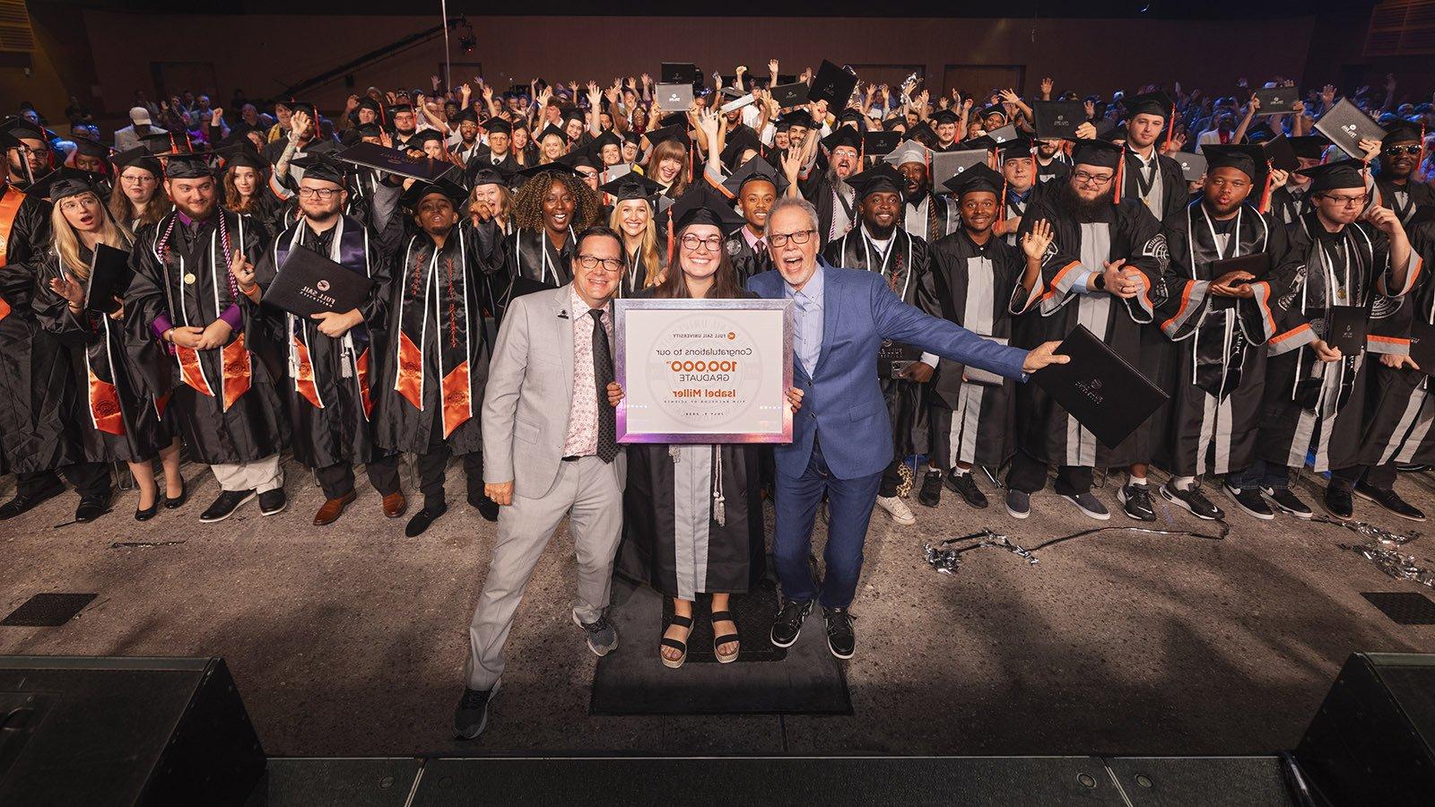 In a large venue full of college graduates, two men in suits stand on opposite sides of a woman in a cap and gown holding a framed photo that reads "满帆 University - Congratulations to our 100,000毕业 Isabel Miller."